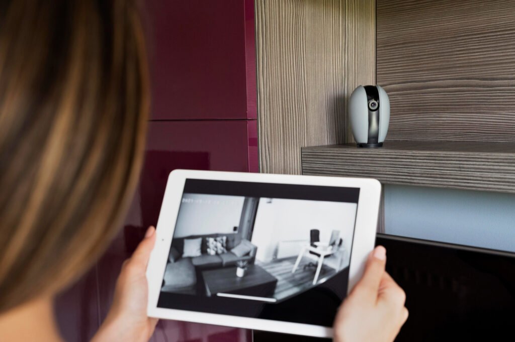 Mulher monitorando sua casa em um tablet com imagens de câmera de segurança, enquanto uma câmera de vigilância real está em uma prateleira, ilustrando um "checklist casa segura".