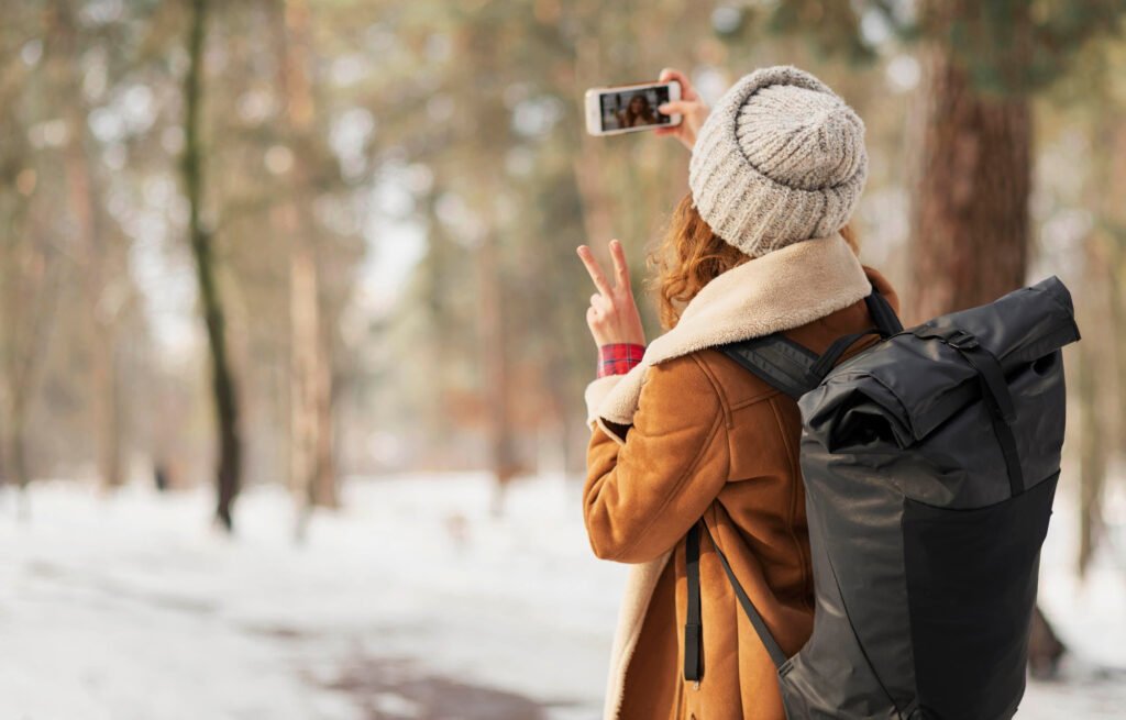 Mulher com mochila e gorro tirando uma selfie com sinal de paz em um cenário de floresta nevada, representando a alegria de viajar e a necessidade de segurança nas férias.